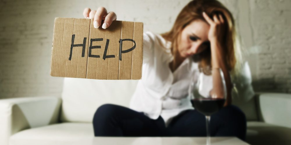 drunk alcoholic blond woman drinking whiskey glass asking for help holding message board depressed wasted and sad at home couch in alcohol abuse and housewife alcoholism