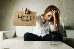 drunk alcoholic blond woman drinking whiskey glass asking for help holding message board depressed wasted and sad at home couch in alcohol abuse and housewife alcoholism
